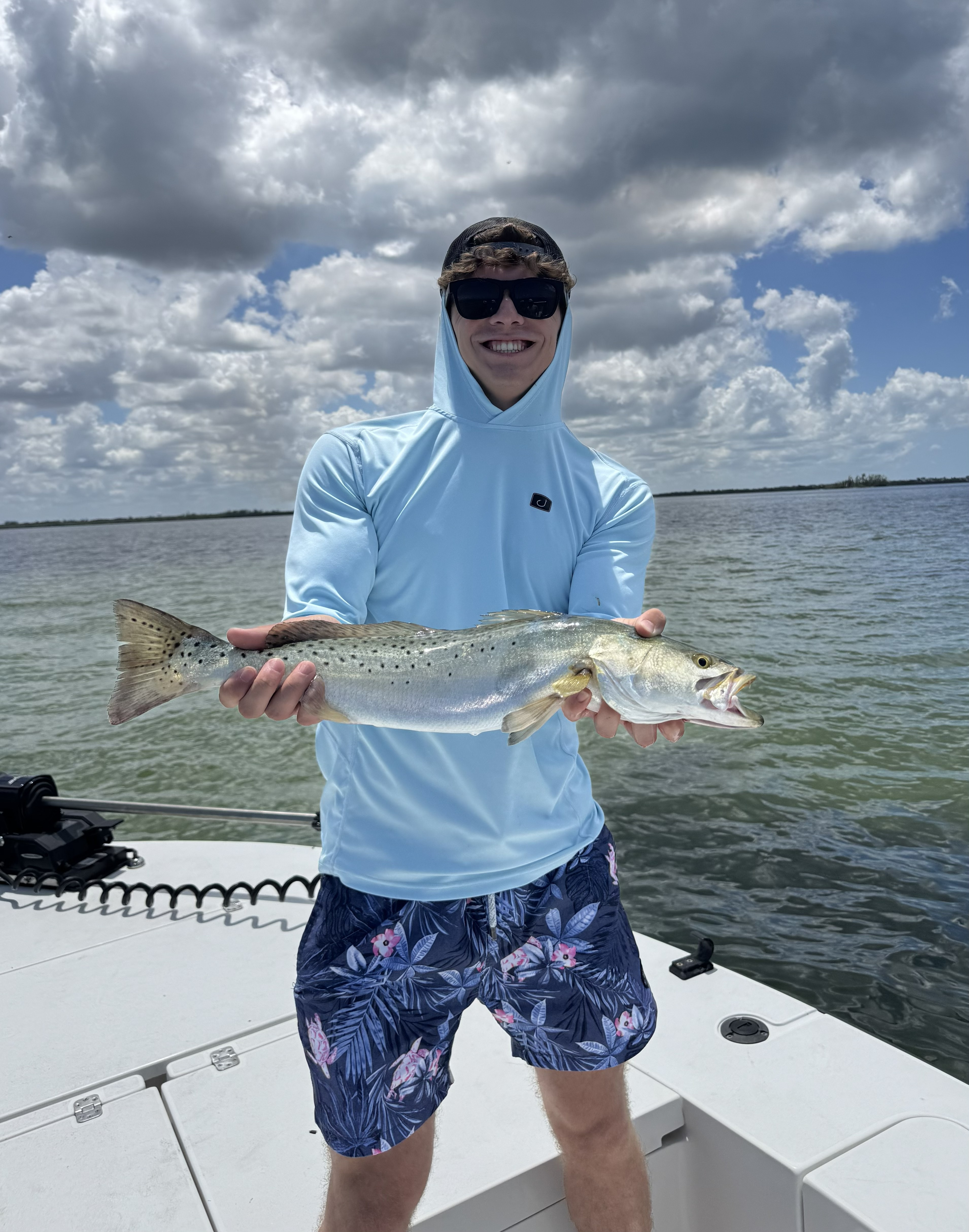 Pierson smiling in sunglasses and a light blue hooded shirt, holding a speckled trout on a boat on a sunny day